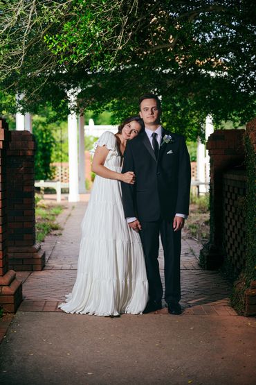 Couple dressed elegantly, woman in white dress leaning on man in black suit under leafy archway.
Wedding Portraiture