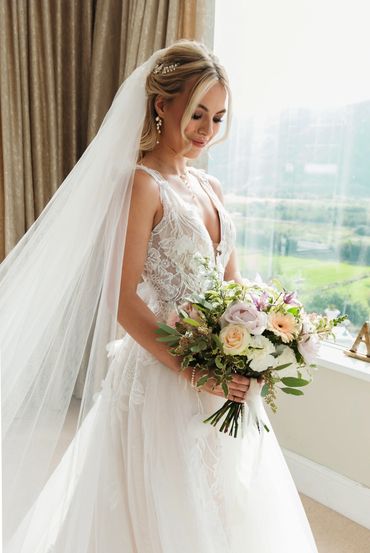 Bride holding light and airy pastel bouquet of fresh flowers in her wedding dress in bridal suite