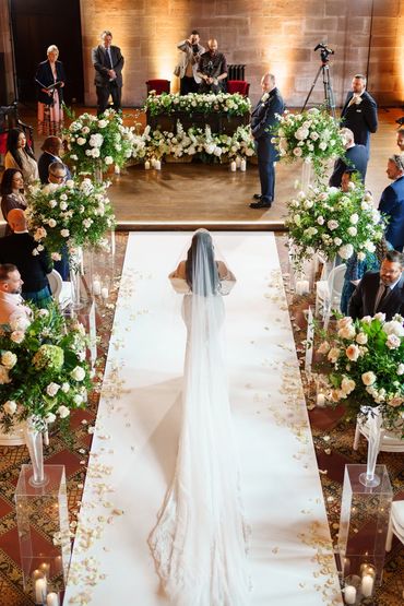 Bride walking down the aisle at Peckforton Castle on white carpet through a meadow of white flowers.