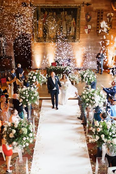 Couple walking down the flower lined aisle at Peckforton Castle with confetti in the air.