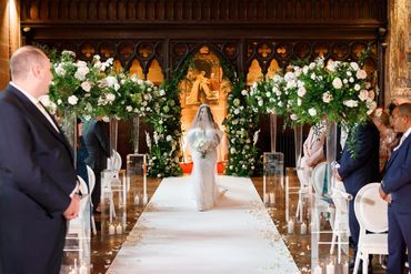 Bride walking down the aisle with flowers and arch in the background in white and nude colours.