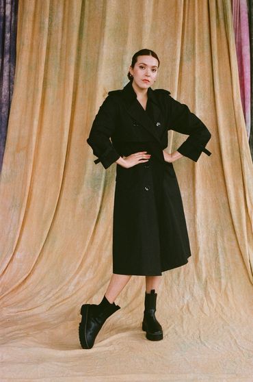 @marissa_cisneross in front of cream tie-dye backdrop, in black trench coat & boots