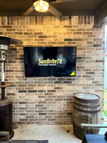 Outdoor TV mounted on a brick wall with a heater and wooden barrel nearby.