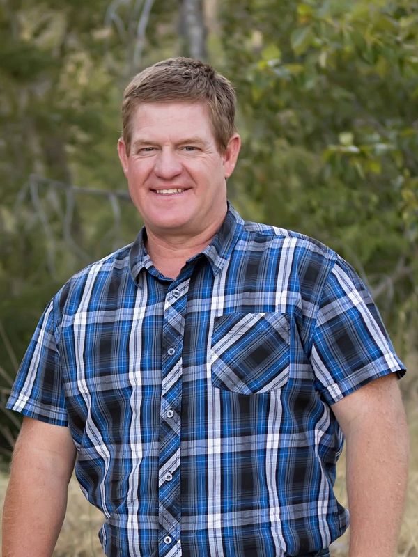 Smiling man in a blue plaid shirt outdoors with greenery background.