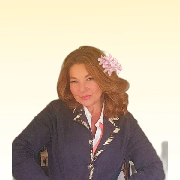 Woman with brown hair and a pink flower in her hair, wearing a dark blazer with patterned trim.