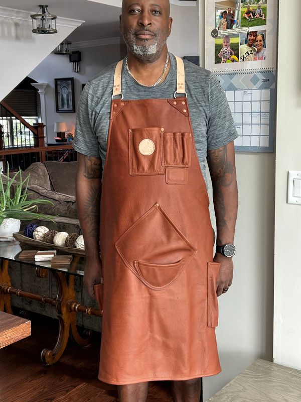 Man wearing a brown leather apron standing indoors near a calendar.