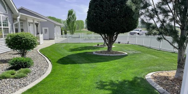 A well-maintained green lawn with trees and a white picket fence around a gray house.