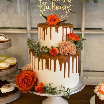 Two-tier white cake with caramel drip and orange flowers on a stand.