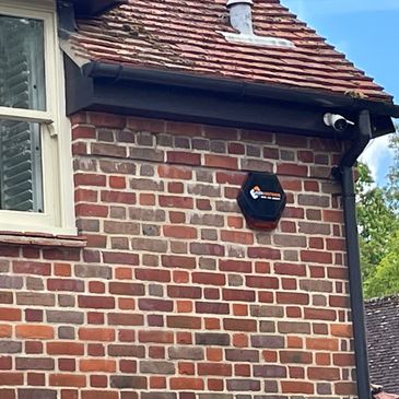 Surveillance camera and alarm system on a brick house wall under a tiled roof.