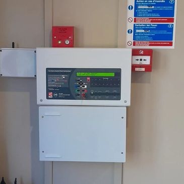 Fire alarm control panel with umbrellas in a stand below it on a beige wall.