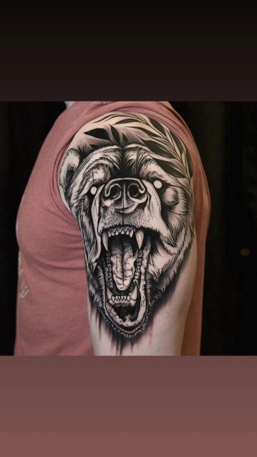 Black and grey realistic roaring bear tattoo on the upper arm, featuring detailed fur, an open mouth, and botanical leaves.