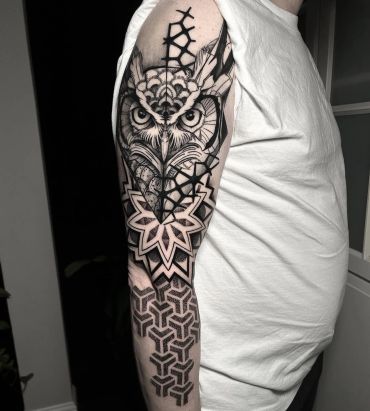 Black and grey owl tattoo on the arm, featuring detailed feathers, bold geometric mandala patterns, intricate linework, and ornamental designs.