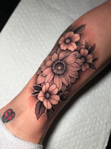Black and grey floral forearm tattoo featuring detailed daisies, layered flower petals, leaves, and soft shading.