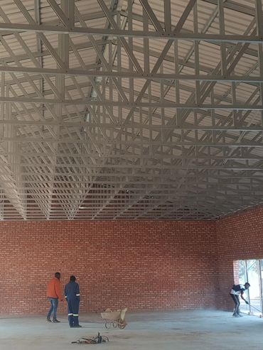 Large brick-walled hall under construction with workers and metal roof trusses.