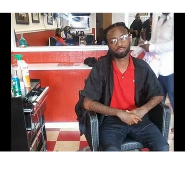 Man sitting in a barber chair wearing glasses and a black cape.