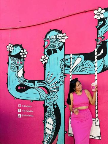 Woman in pink outfit holding a drink, posing by colorful cactus mural on pink wall.