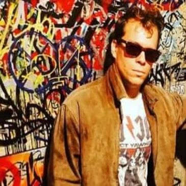 Man wearing sunglasses and a brown jacket stands in front of a colorful graffiti wall.