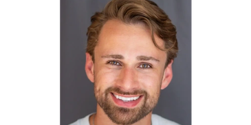Smiling young man with light brown hair and beard against a gray background.
