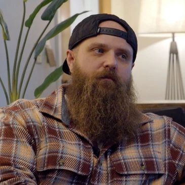 Bearded man in a plaid shirt and backward cap indoors.