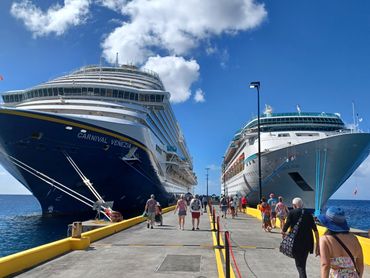 People walking on a pier between two docked cruise ships under a sunny sky.