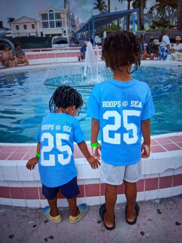 Two children in matching blue "Hoops @ Sea 25" shirts holding hands near a water fountain.