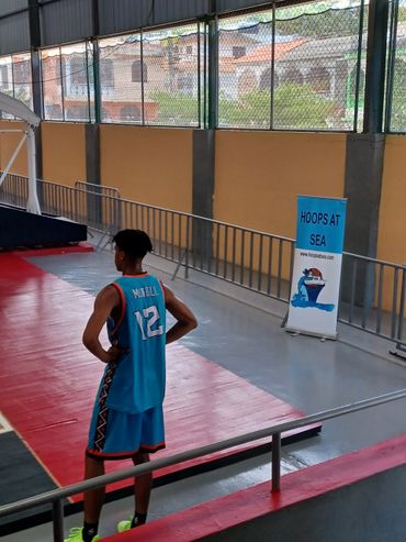 Basketball player in blue jersey standing on indoor court near 'Hoops at Sea' banner.