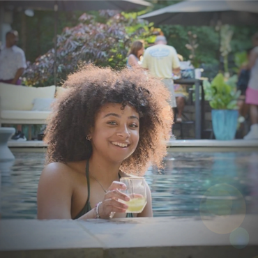 Young woman smiling in a pool holding a drink, enjoying a sunny day.