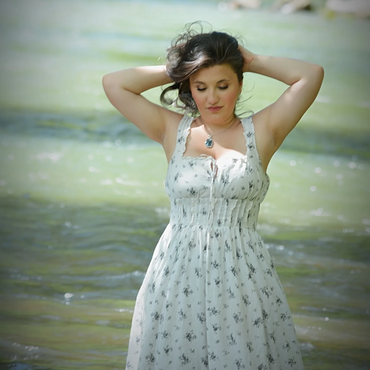 Woman in a white floral dress standing in a river with hands behind her head.