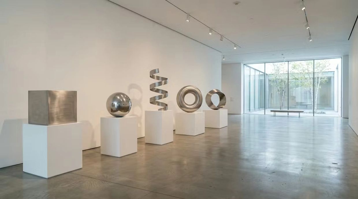 Modern metal sculptures displayed on white pedestals in a minimalist gallery.