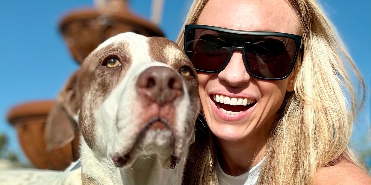 Woman with sunglasses smiling next to a large dog outdoors on a sunny day.