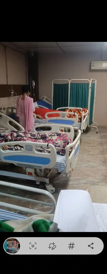 A hospital room with several beds and patients, and a woman in pink standing beside one bed.