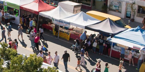 People walk and shop at outdoor market stalls with colorful canopies.