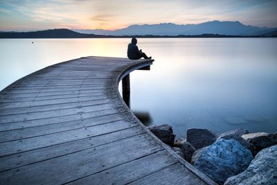 Person sitting alone on a curved wooden pier by calm water at sunset.