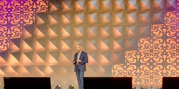 Man in blazer speaking on stage with decorative geometric backdrop.