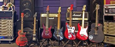 7 guitars lined up in front of power amplifiers, speakers, and Marshall amps