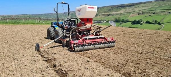 Photograph of a small tractor pulling a seed drill in a field.