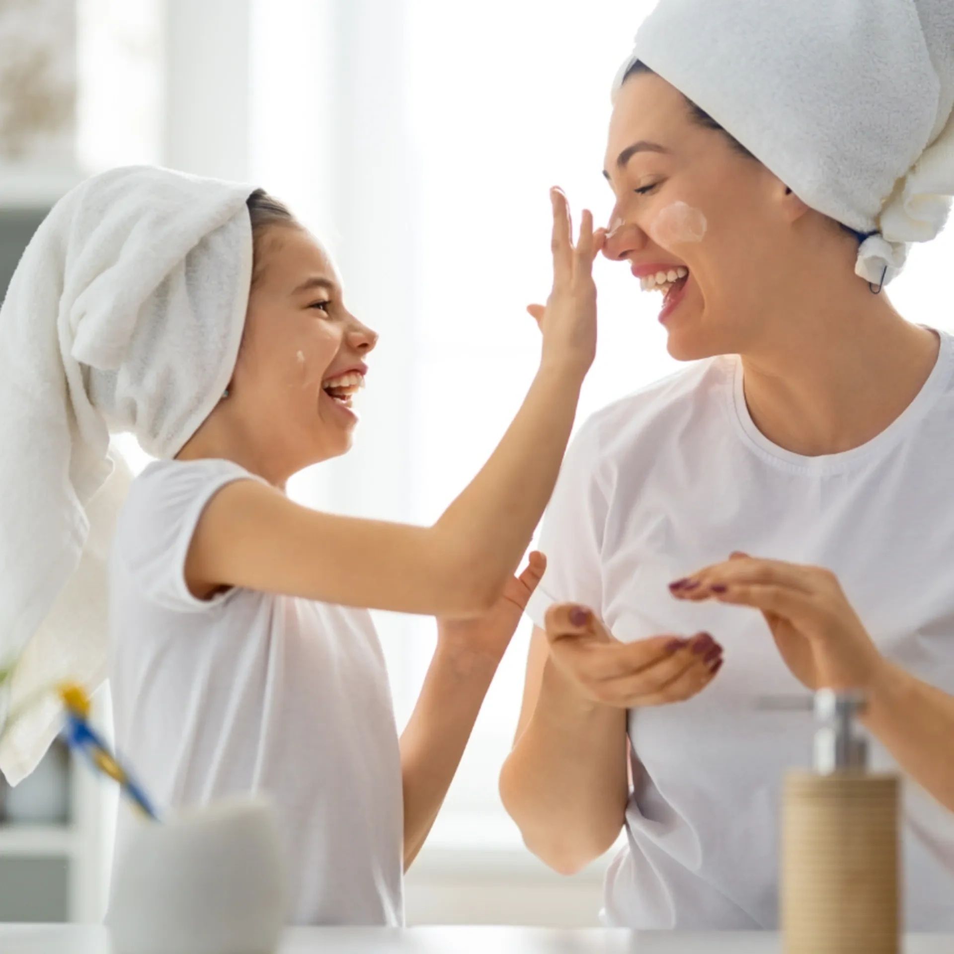 Small girl putting lotion on her mom's nose. Both have towels on their heads and are laughing.
