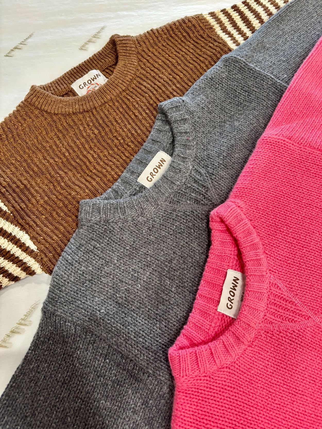 Three cozy knit sweaters in brown, grey, and pink from the brand GROWN.