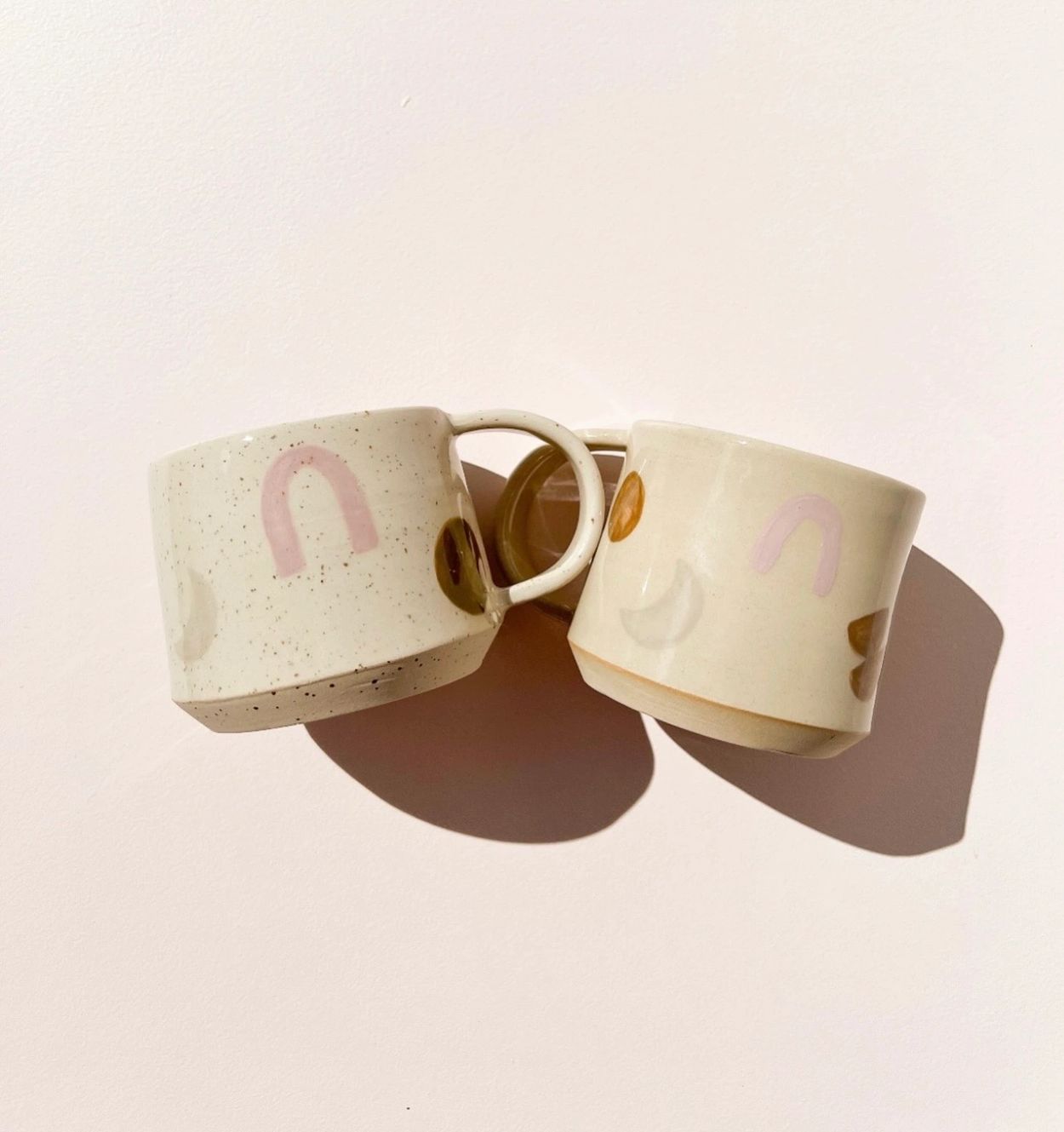 Two ceramic mugs with pastel designs casting shadows on a light surface.
