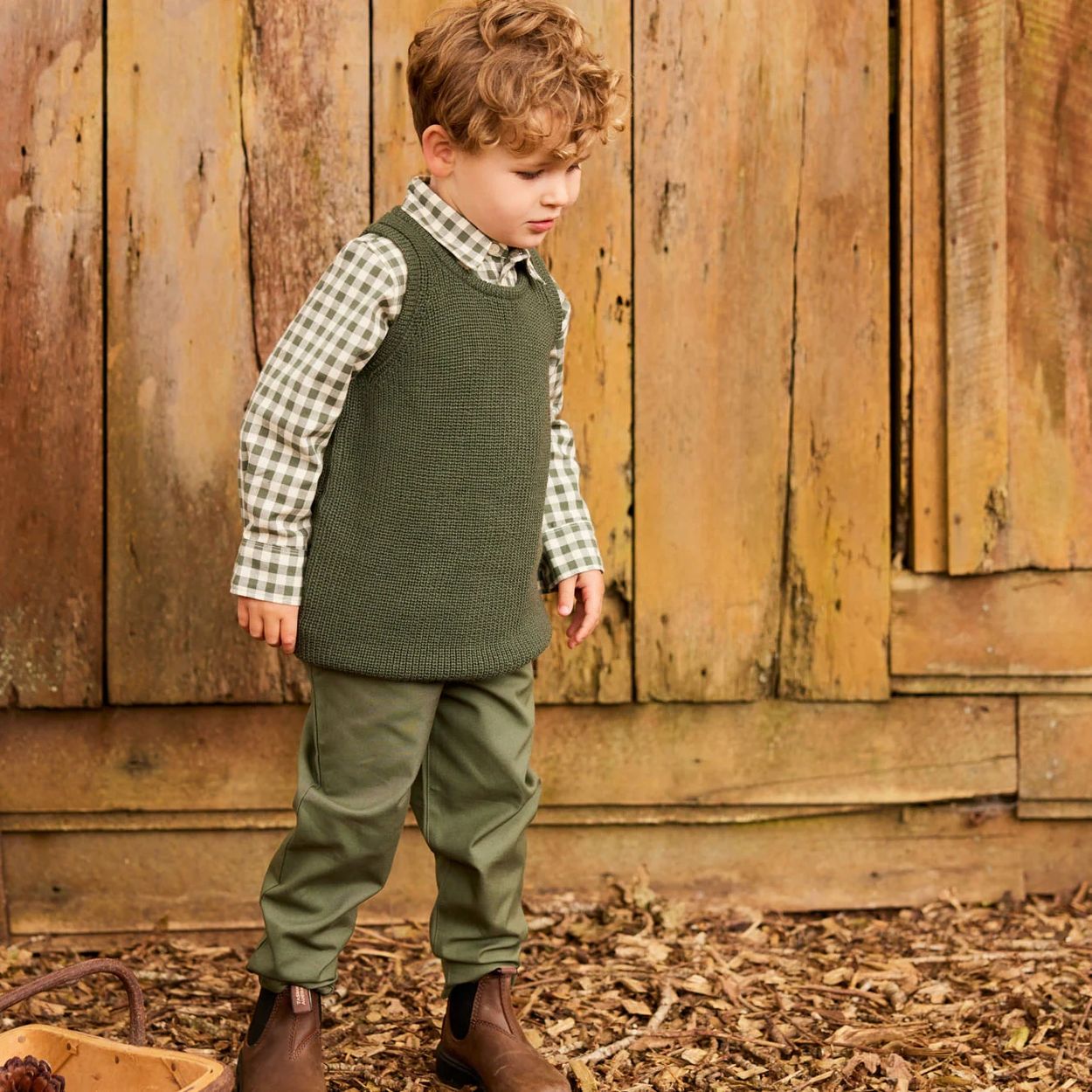 A young boy in green outfit standing near a rustic wooden wall.