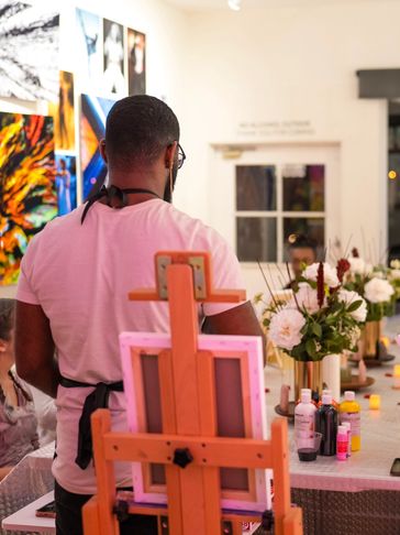 A group art class with people painting on canvases at a long table.