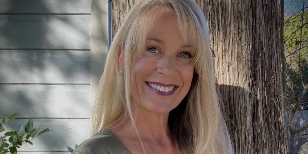 Smiling woman with long blonde hair outdoors near a tree.