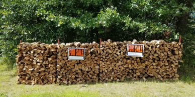 Stacks of firewood for sale with signs in front of a wooded area.