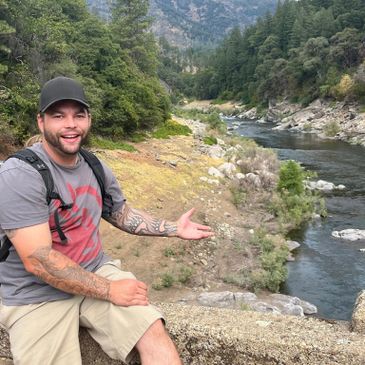 Man with tattoos enjoying a scenic river and forest view.
