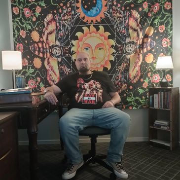 Man seated in front of a vibrant sun and moon tapestry in a cozy room.