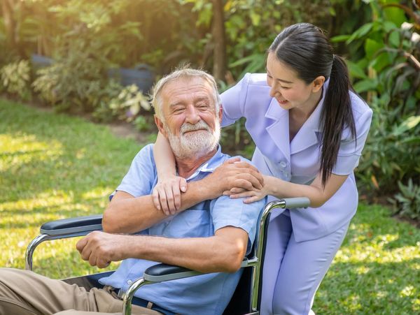 Nurse warmly embraces elderly man in wheelchair outdoors.