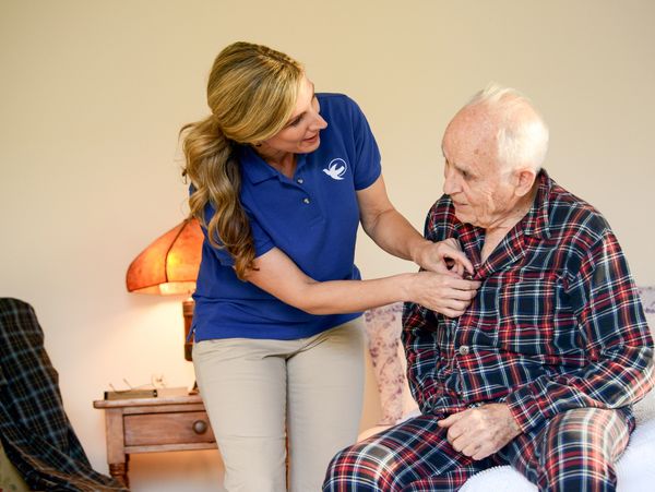 A caregiver helps an elderly man button his pajamas in a cozy room.