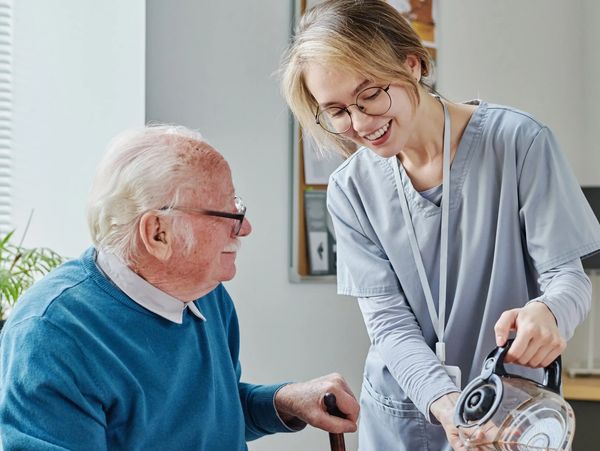 A nurse smiling while pouring a drink for an elderly man.