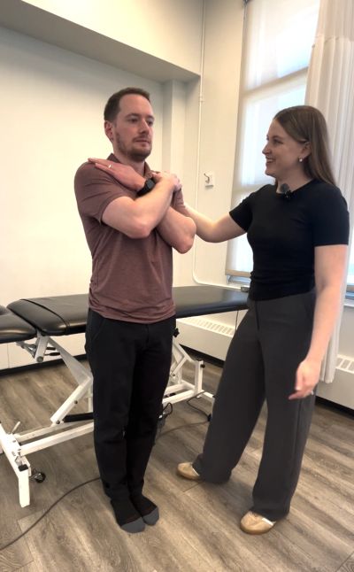 A man performs a neck stretch with guidance from a woman in a clinical setting.