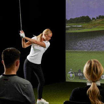 Woman playing golf on an indoor golf simulator with two people watching.